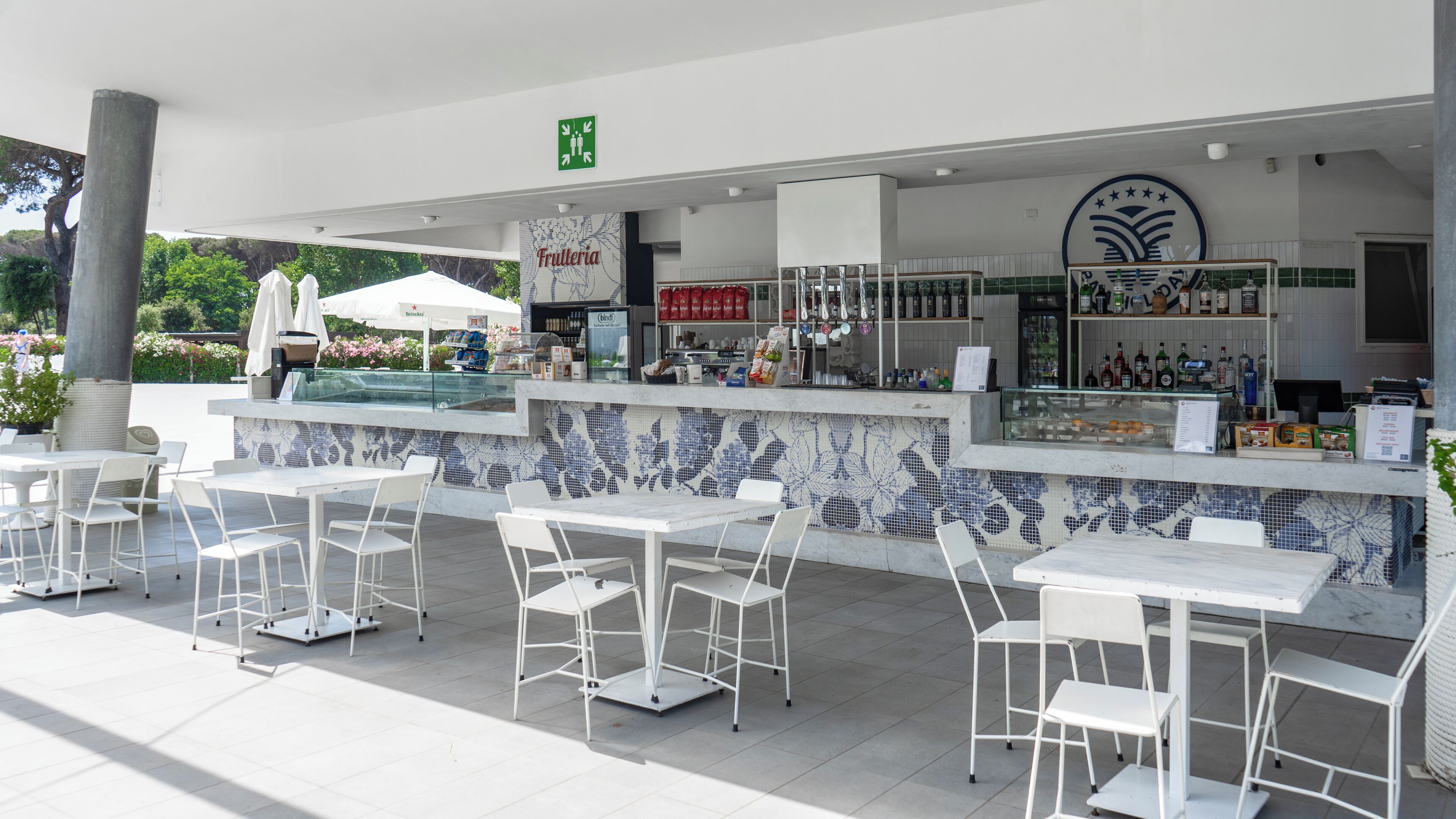 Zona de bar al aire libre con mesas y sillas blancas, un revoque de azulejos y una variedad de botellas y aperitivos en estantes.