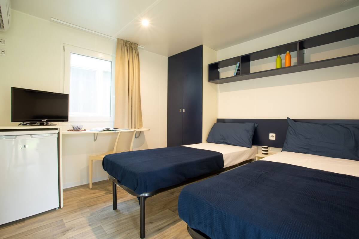 A hotel room with two beds, a desk, a television, and a small kitchenette.