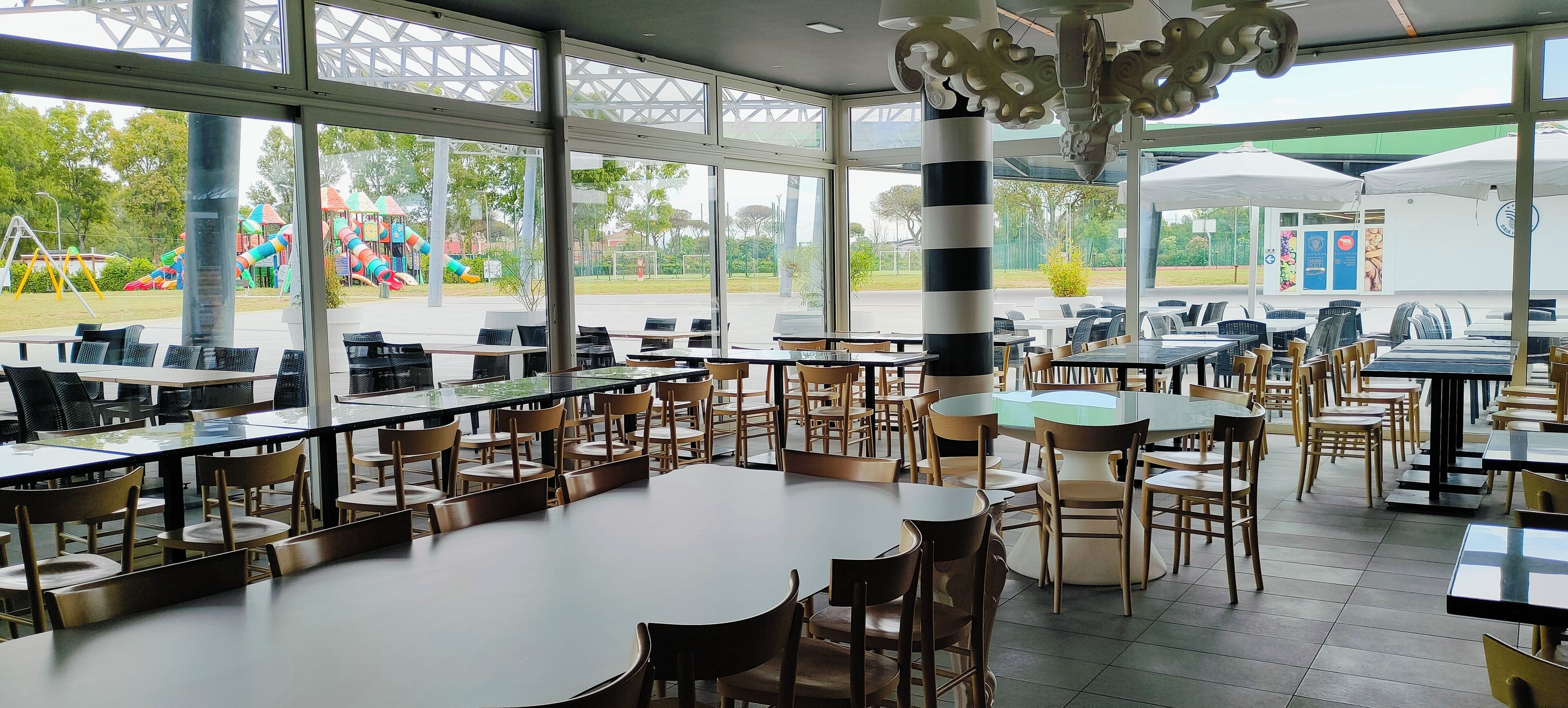 A spacious indoor dining area with large windows, multiple tables and chairs, and a view of an outdoor playground.