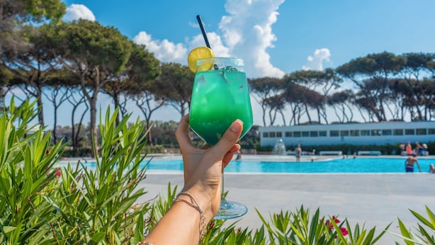 A hand holding a green drink with a lemon slice and straw, in front of a swimming pool with trees and a building in the background. - Camping Village Roma Capitol (Rome)