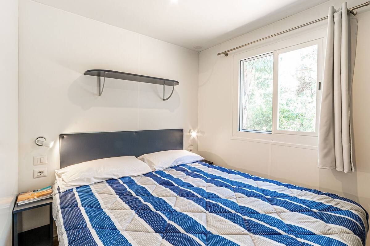 A bedroom with a bed, a nightstand, a wall-mounted shelf, a window with curtains, and a view of greenery outside.
