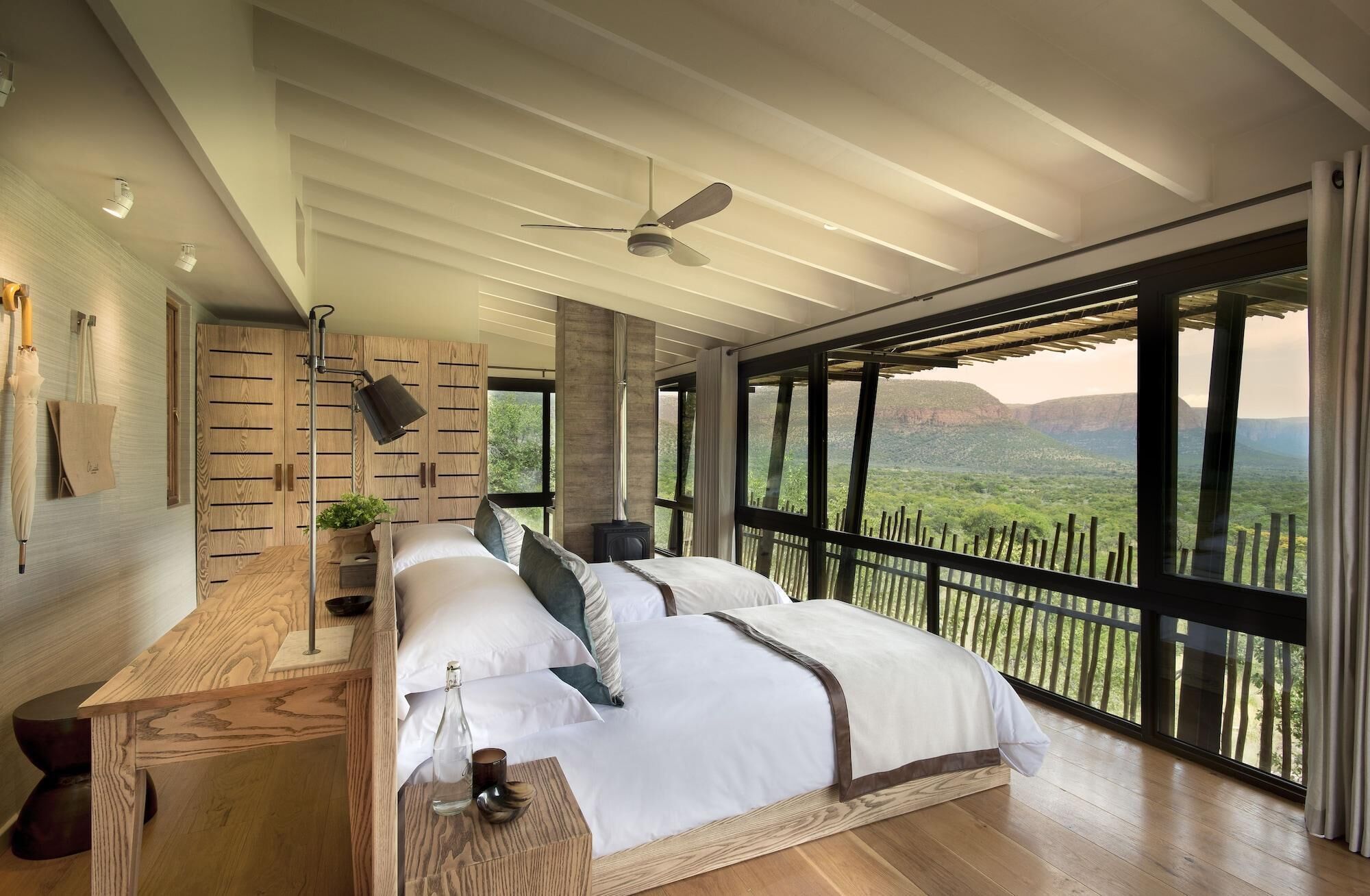 A bedroom with a large bed, a ceiling fan, a wooden wardrobe, and a view of a mountainous landscape.