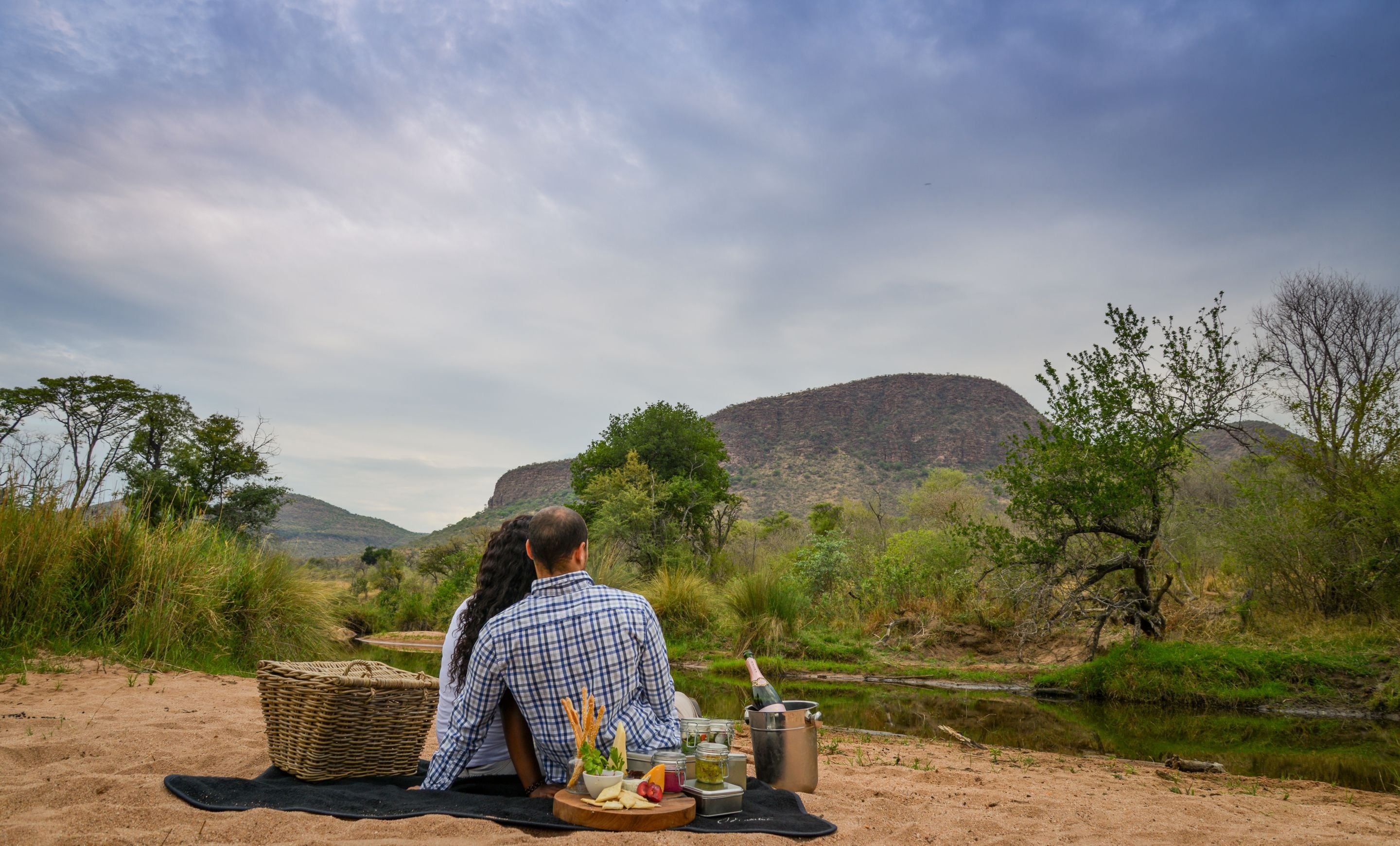 Een stel zit op een deken langs een rivier, met een picknickopstelling inclusief een mand, fles en eten.