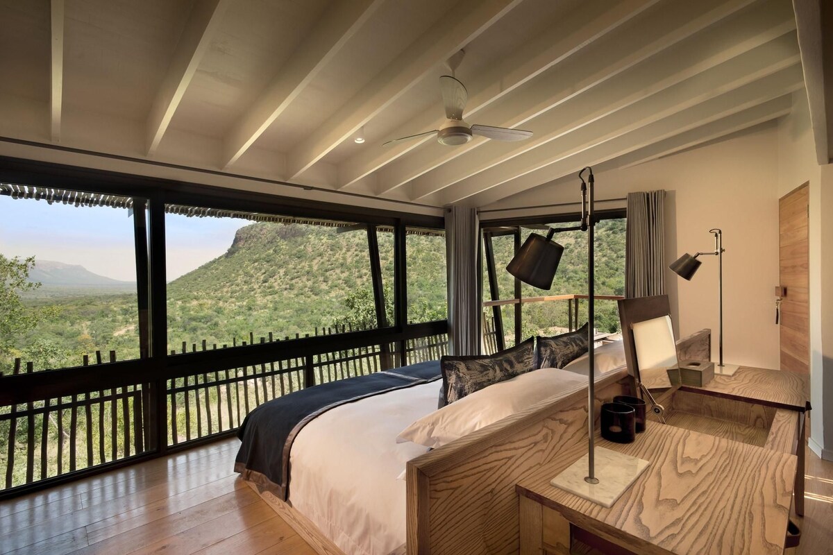 A bedroom with a large bed, a desk, a chair, a lamp, and a view of a mountainous landscape.