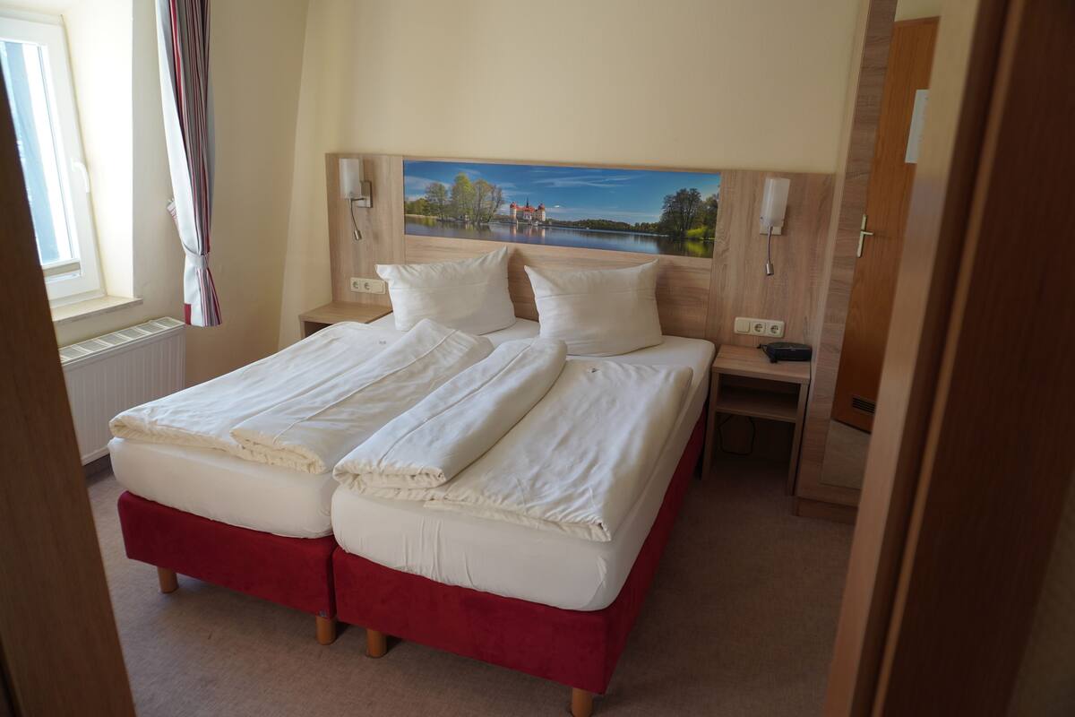 A double bed with white linens, a red upholstered footboard, and a headboard with a landscape painting.