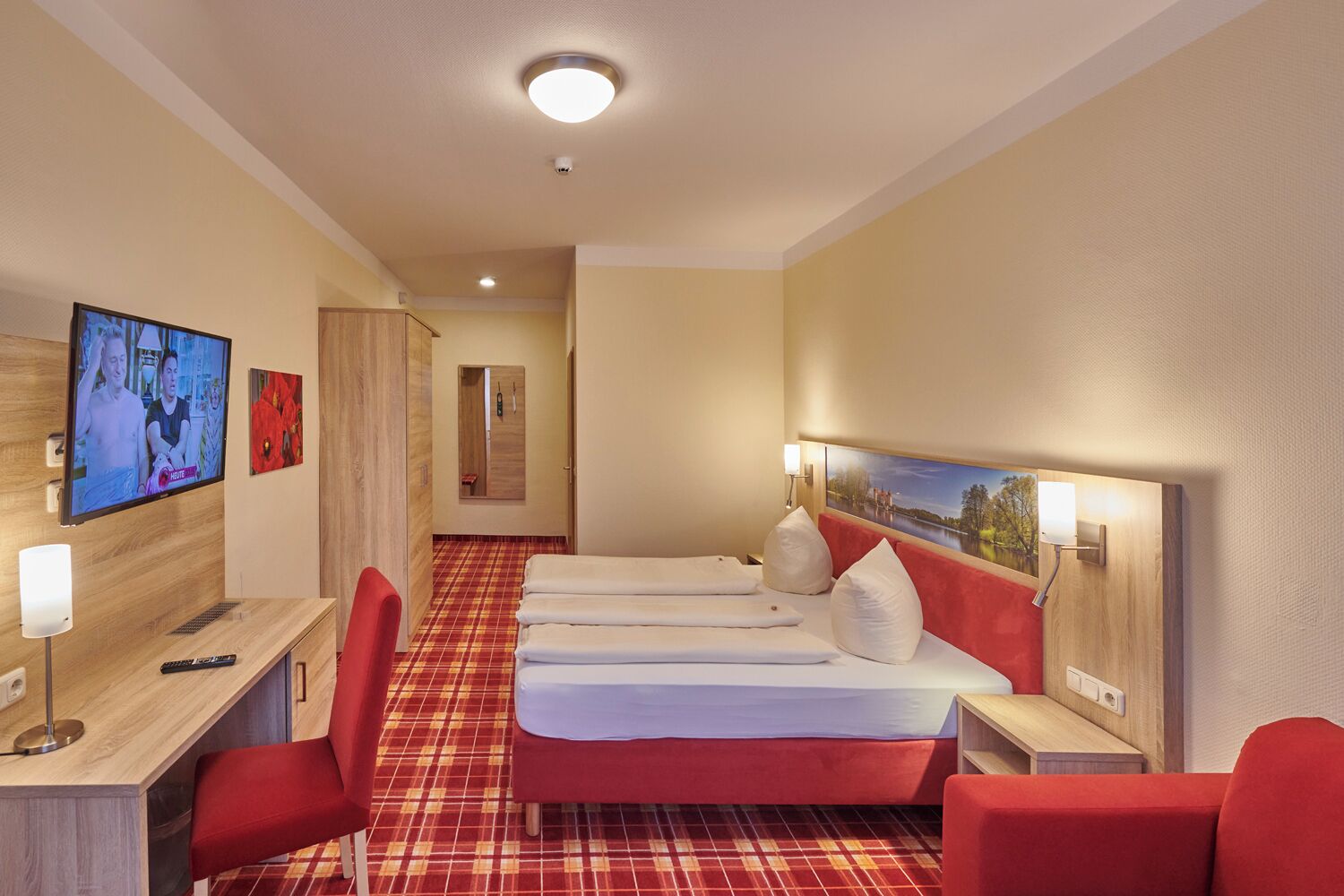 Quarto de hotel com chão xadrez vermelho, cama com roupa de cama branca, escrivaninha com computador, televisão na parede e um quadro sobre a cama.