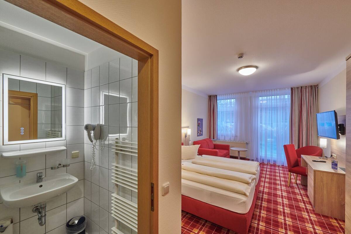A hotel room with a bathroom, a bed, a desk, a TV, and a window with curtains.
