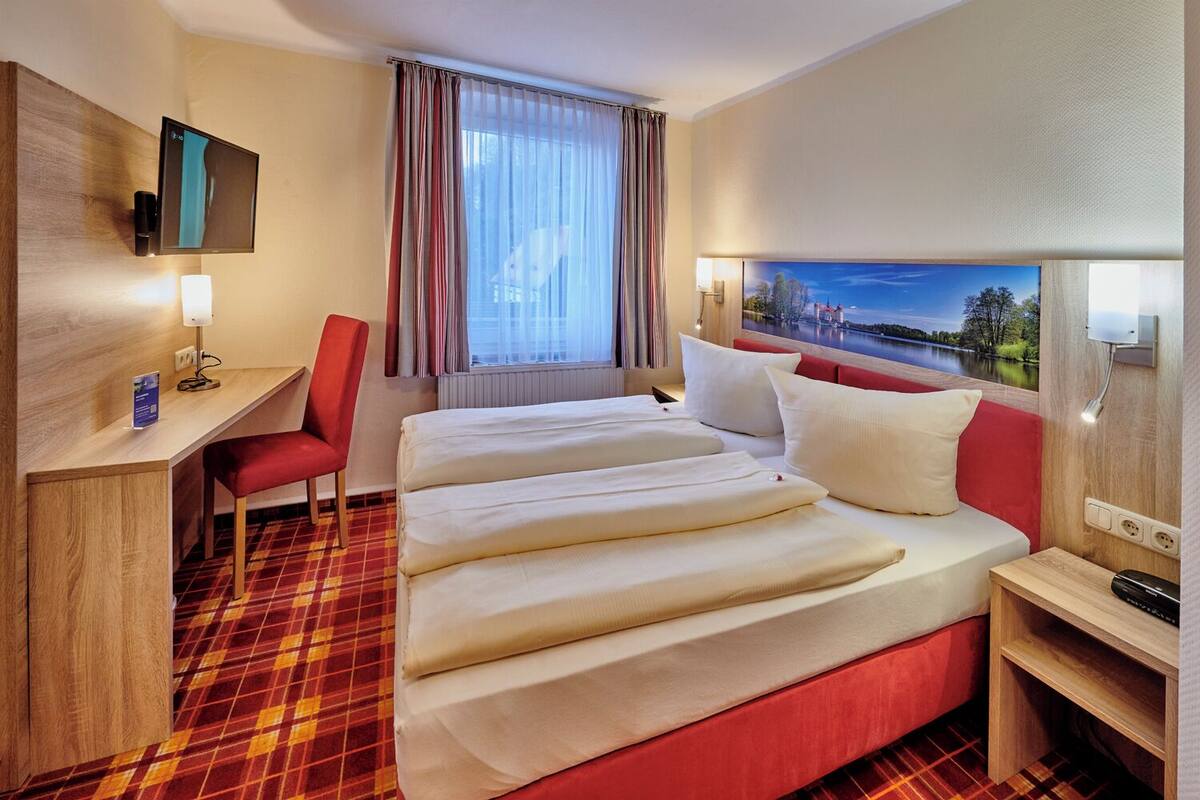 A hotel room with a bed, a desk, a chair, a TV, and a window with curtains.