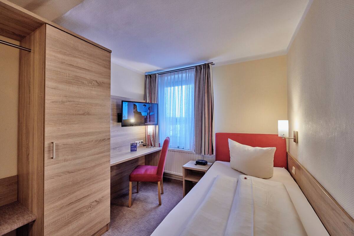 A hotel room with a wooden wardrobe, a desk with a chair, a bed with a red headboard, and a television on the wall.