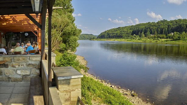 A riverside restaurant with outdoor seating, overlooking a calm river and forested hills. - Hotel Elbparadies (Pirna)