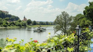 A river with a boat, greenery, and a building in the background. - Hotel Elbparadies (Pirna)