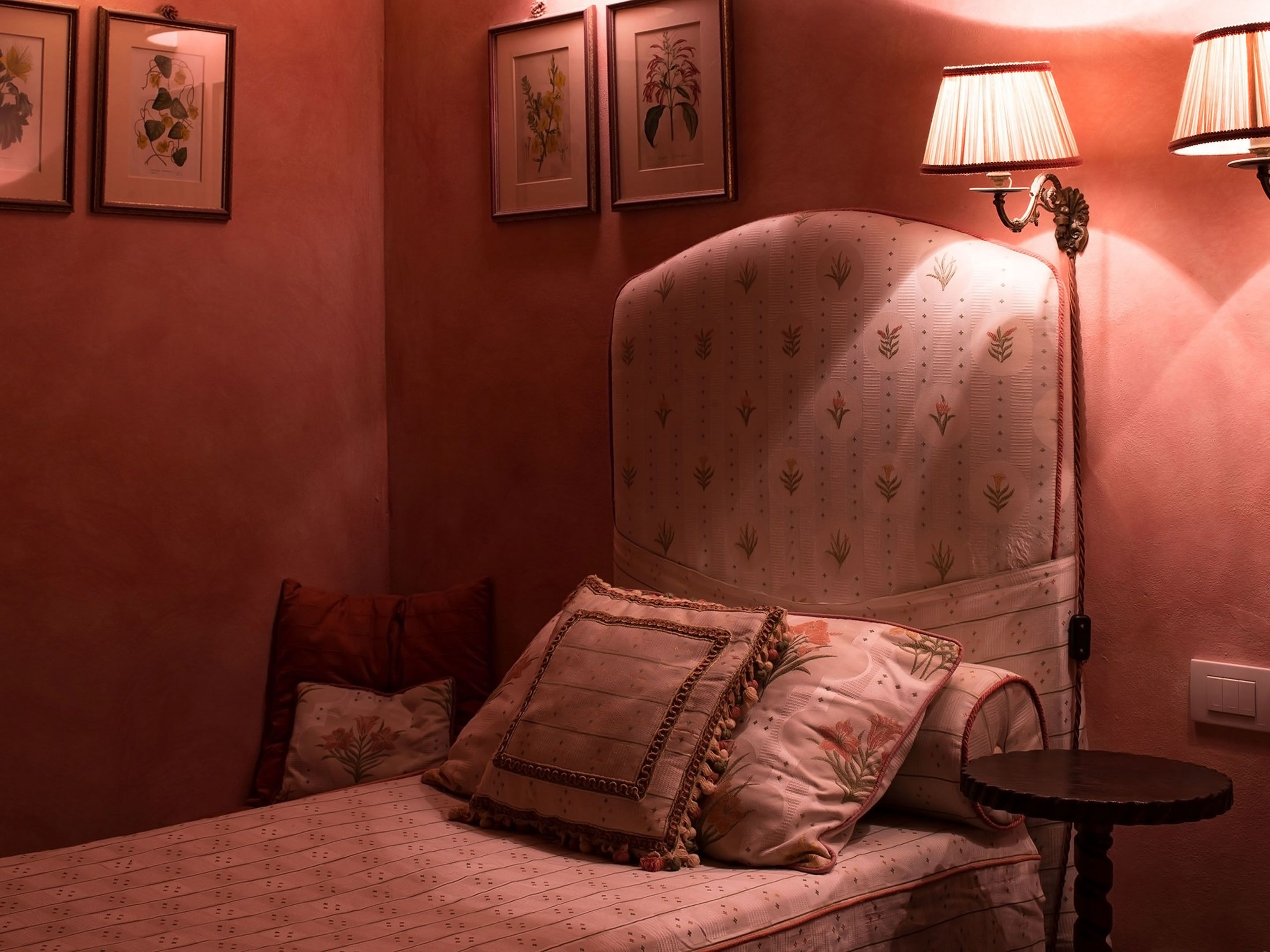 A bedroom with a bed, pillows, a bedside table, a lamp, and framed botanical prints on the wall.