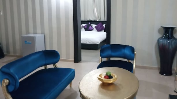 A room with a bed, two blue sofas, a round table with a bowl of fruit, a vase, and a small cabinet. - The Nook Apartments (Abuja)