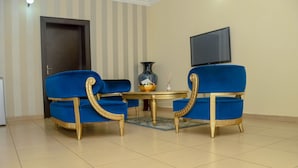 A room with two blue chairs with gold accents, a small round table, a vase, and a television on the wall. - The Nook Apartments (Abuja)
