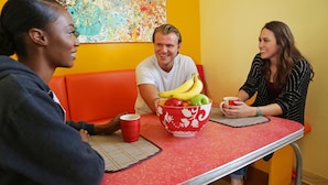 Trois personnes sont assises autour d’une table rouge, devant un saladier de fruits et des tasses rouges.