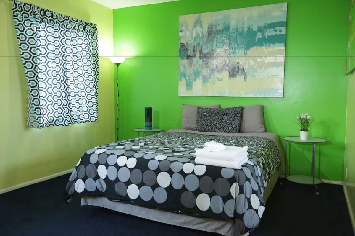 A hotel room with a green wall, a bed with a polka dot bedspread, a painting, a lamp, and a small table with a plant.