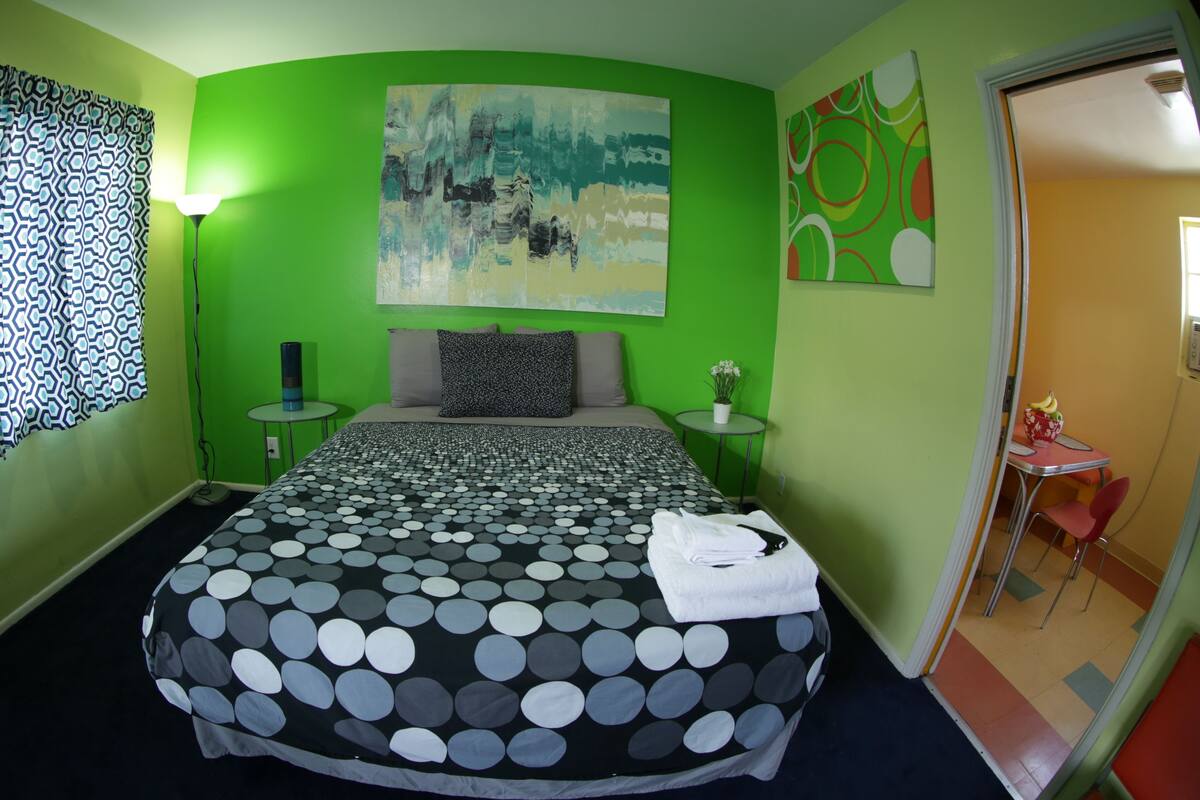 A room with a bed, a small table with a plant, a painting on the wall, and a view of another room.