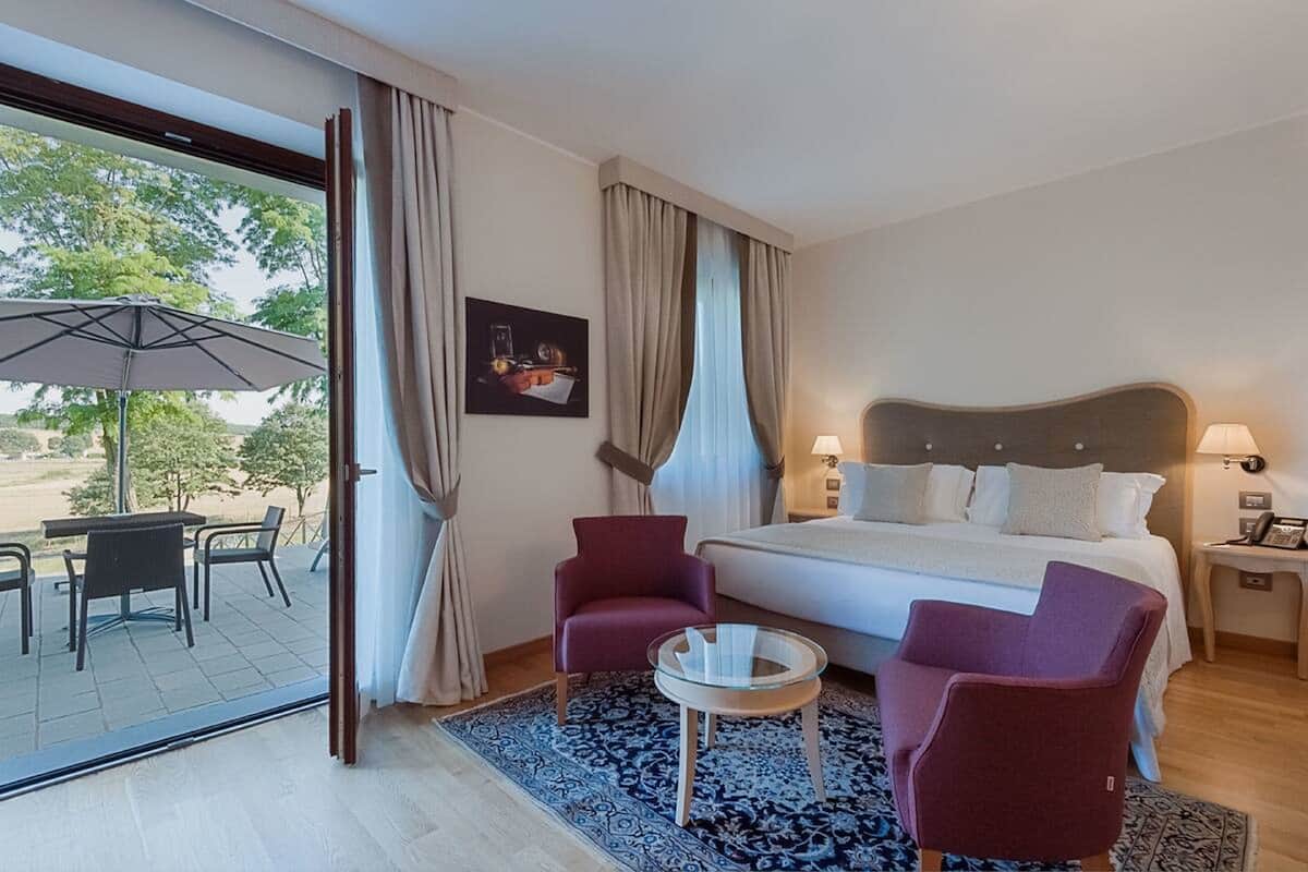 A hotel room with a bed, two armchairs, a small table, a window with curtains, and a view of the outdoors.