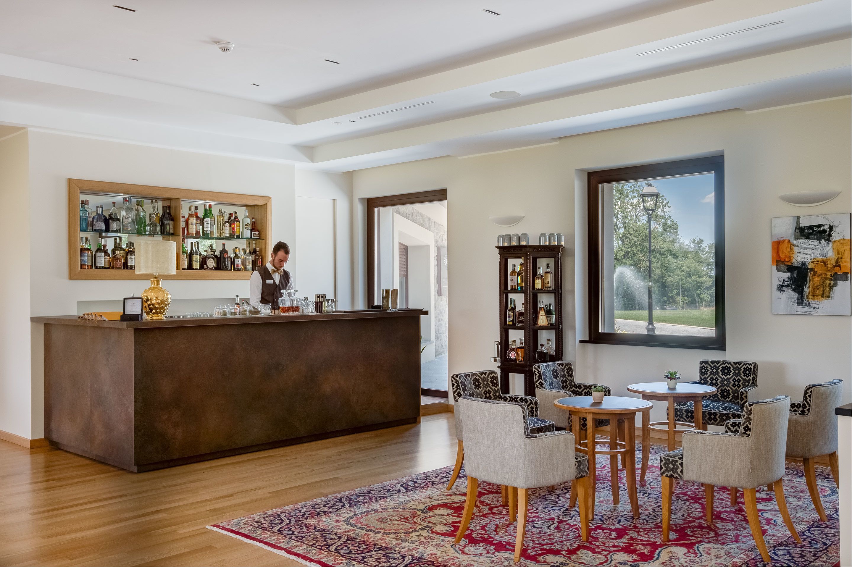 A bar area with a bartender, a large mirror with liquor bottles, and a view of the outdoors through a window.