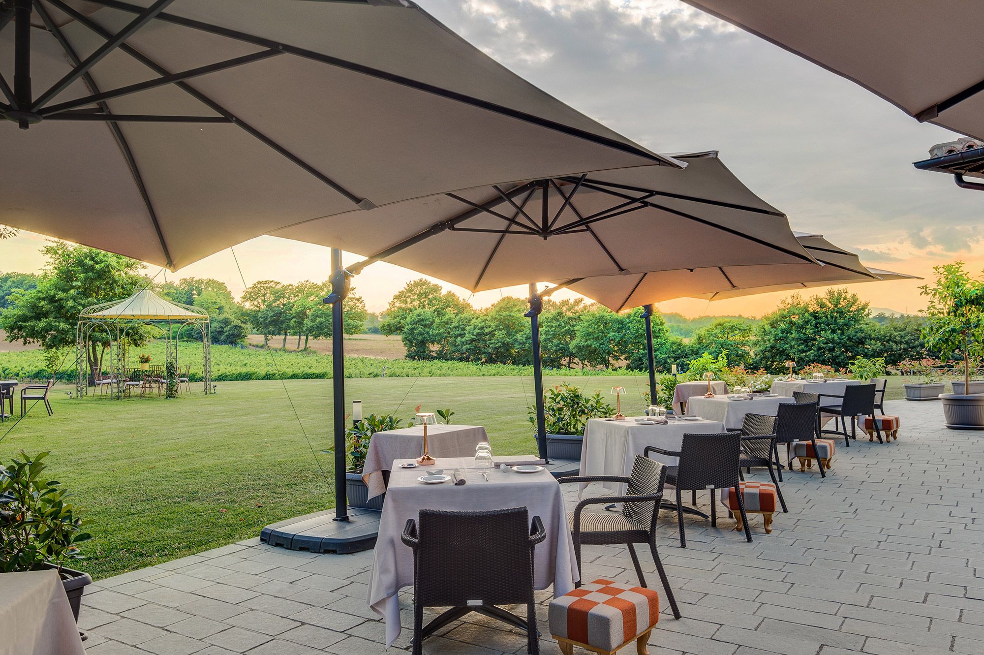 An outdoor dining area with tables and chairs under large umbrellas, set against a backdrop of a grassy field and trees.