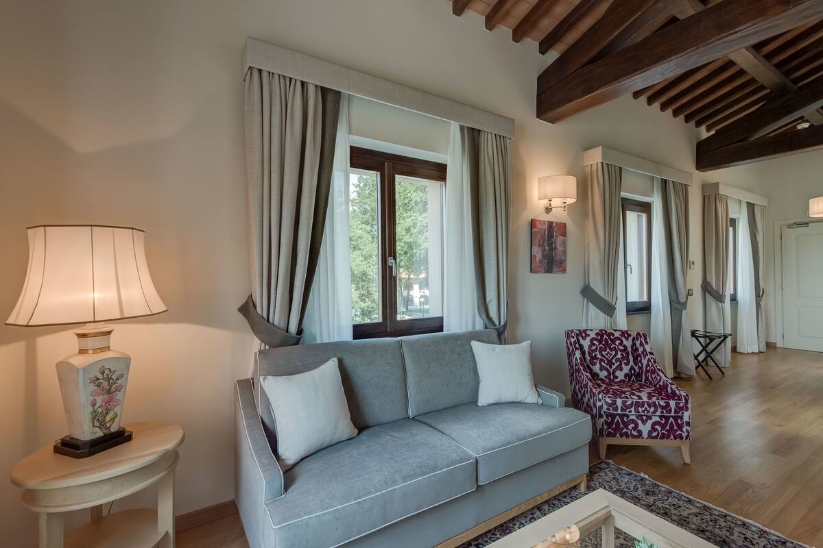 A living room with a grey sofa, a patterned armchair, a lamp, and a window with curtains.