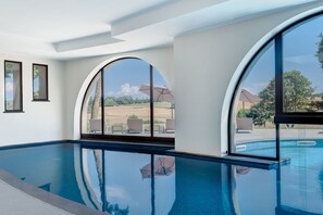 A modern indoor swimming pool with large arched windows, clear blue water, and a view of a landscaped area with outdoor seating.
