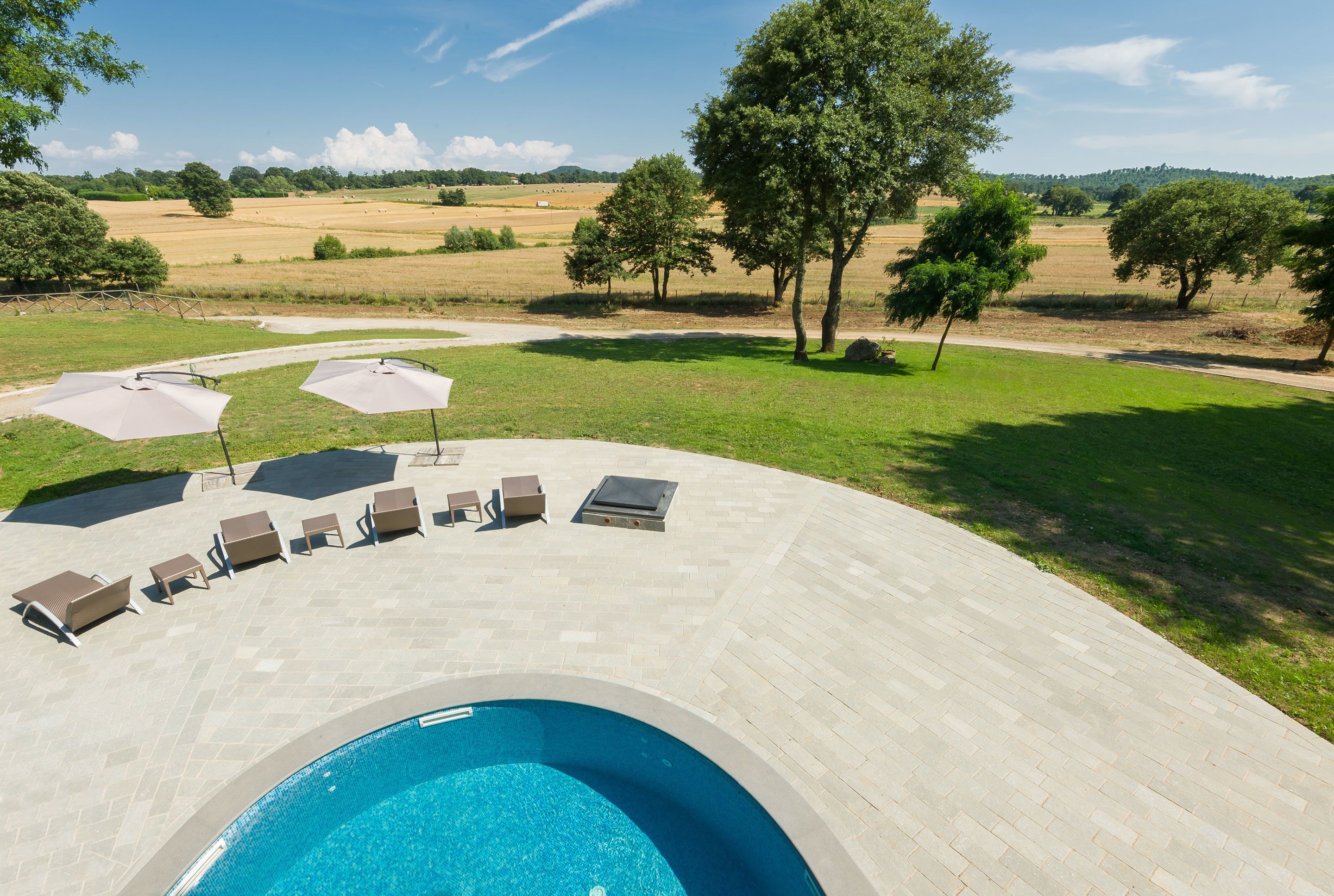 Ein Schwimmbecken mit Liegestühlen und Sonnenschirmen, umgeben von einer Betonplattform und Blick auf eine ländliche Landschaft mit Bäumen und Feldern.
