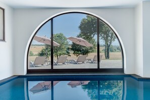 Vista a través de un amplio ventanal de una zona de piscina con sillones de descanso y sombrillas, reflejando la superficie del agua.