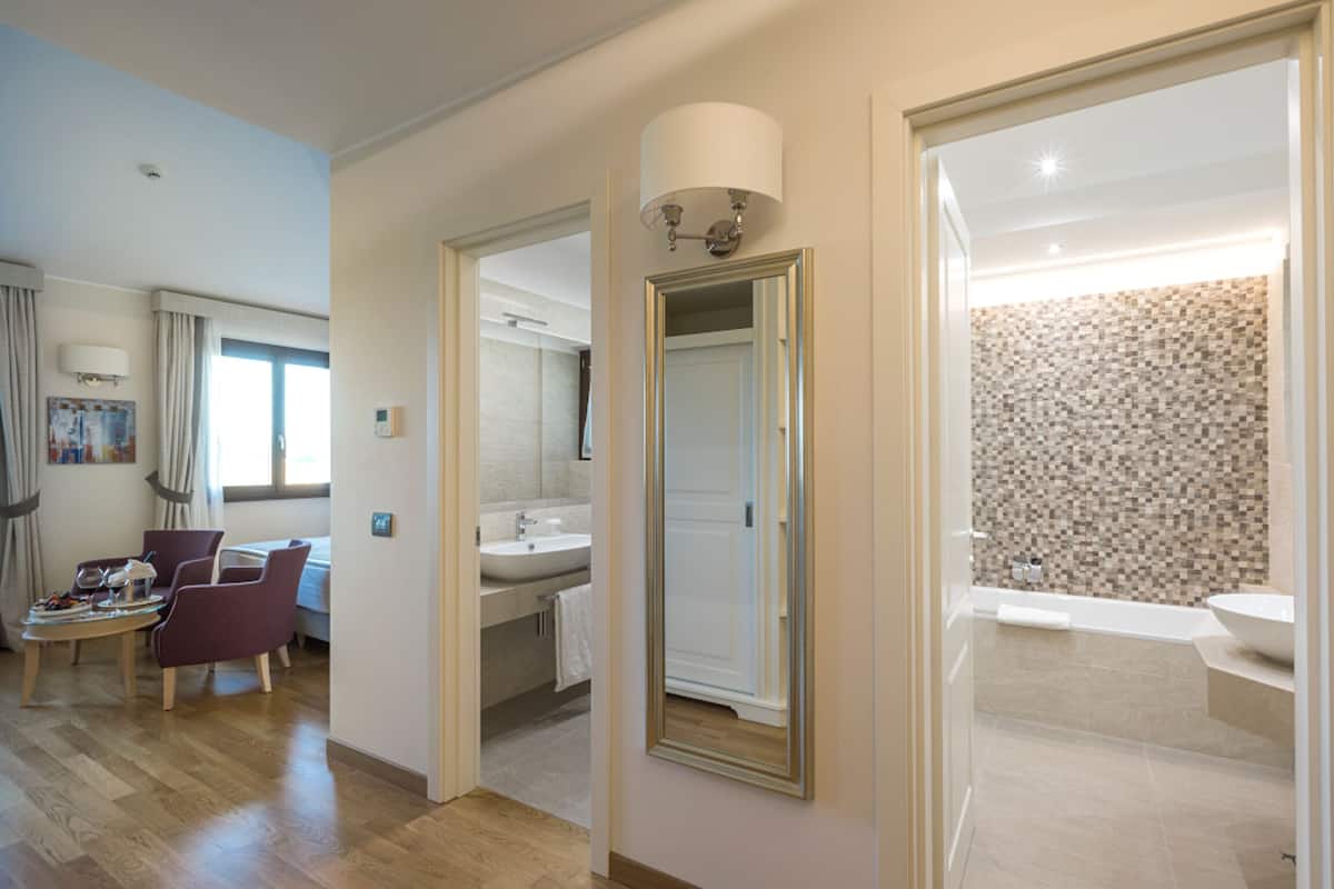 A hotel room with a wooden floor, a sitting area with a table and chairs, a bathroom with a sink and mirror, and a shower area with a tiled wall.