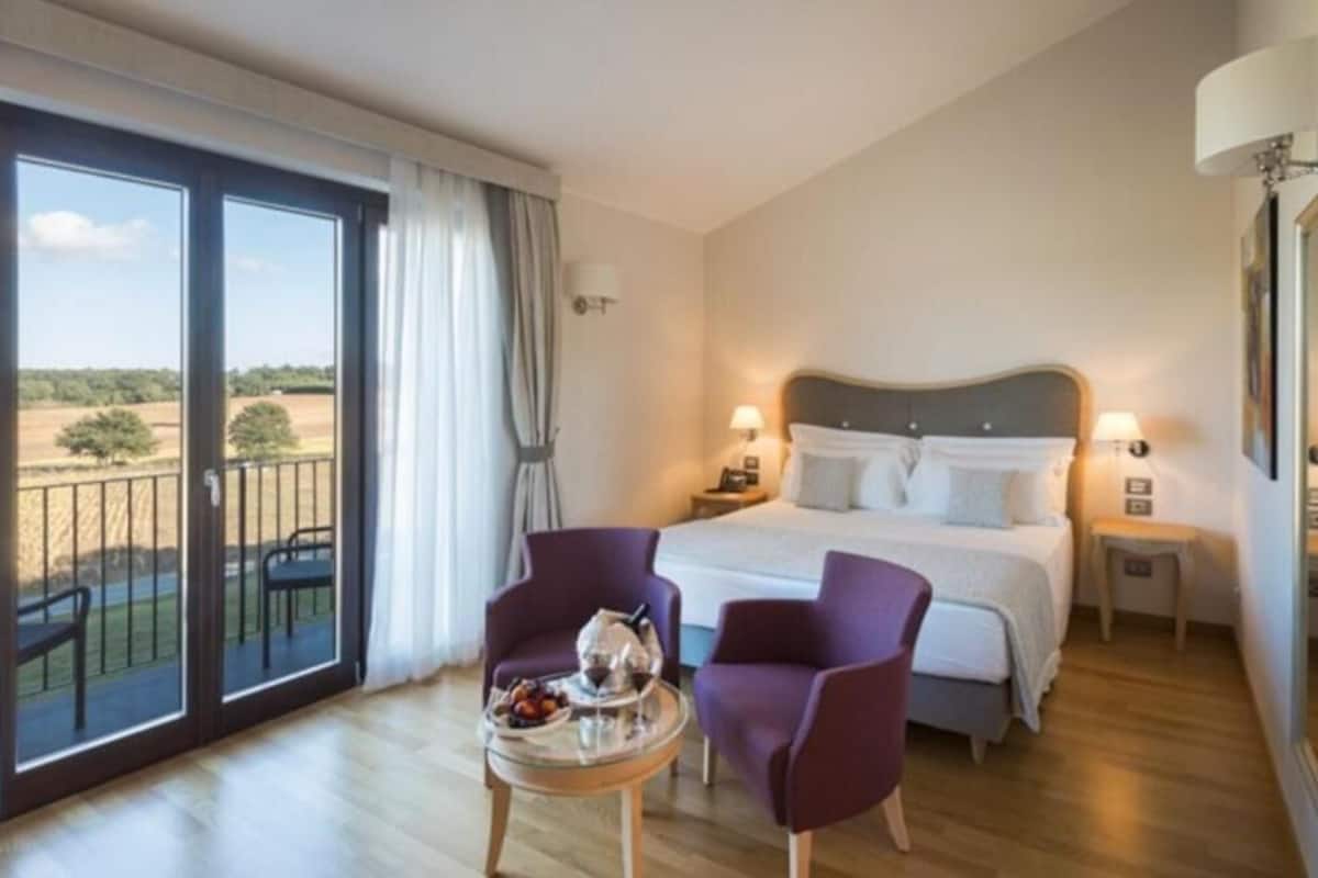 A hotel room with a large bed, two bedside lamps, a small table with a teapot, and a view of the countryside through a sliding glass door.