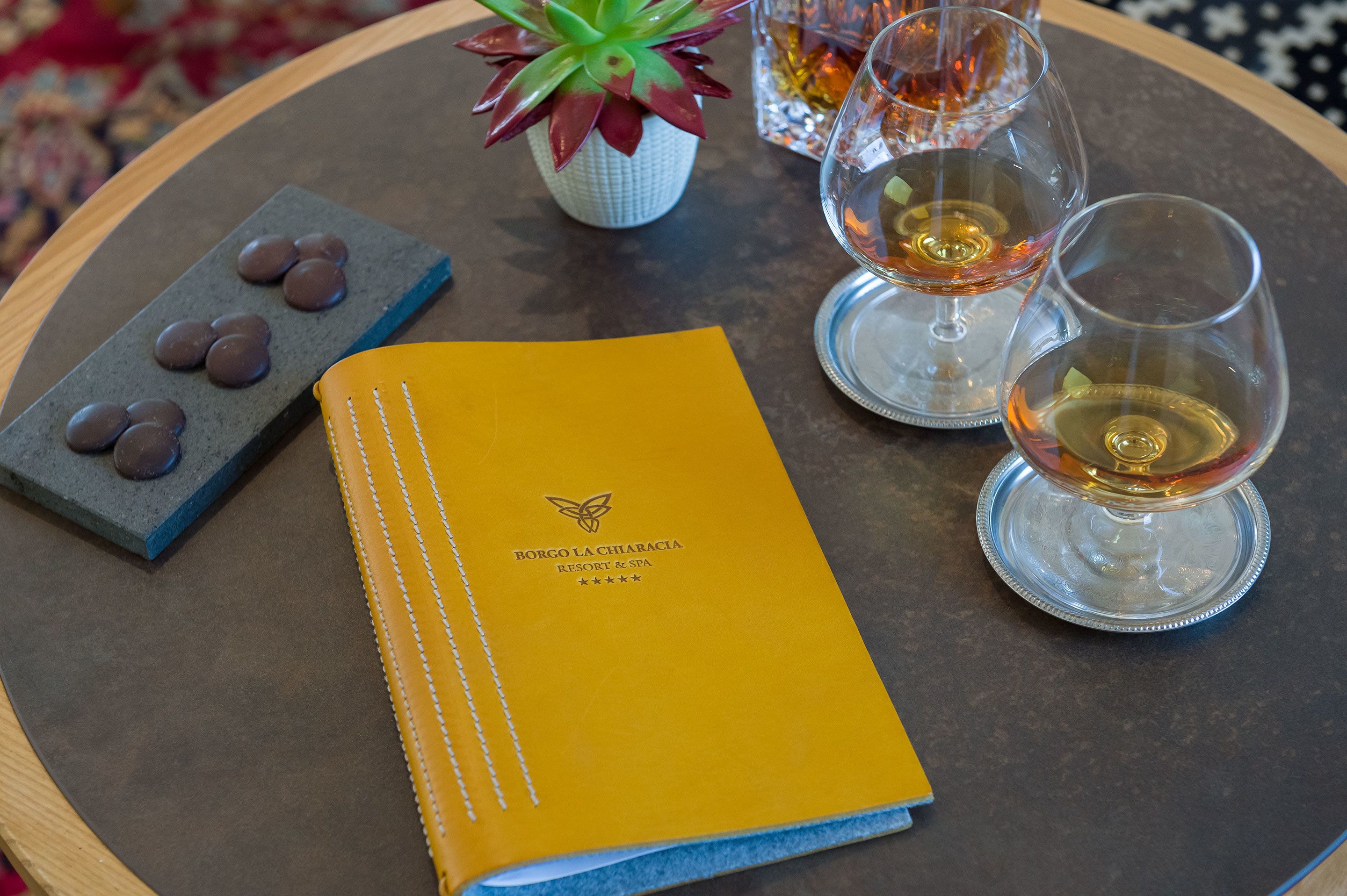 Una mesa con un folleto amarillo, dos copas de vino, una planta pequeña y una bandeja con trufas de chocolate.