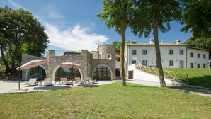Un bâtiment à la façade en pierre, doté d’une grande entrée en arc de cercle et d’une cour verdoyante aménagée avec des chaises longues.