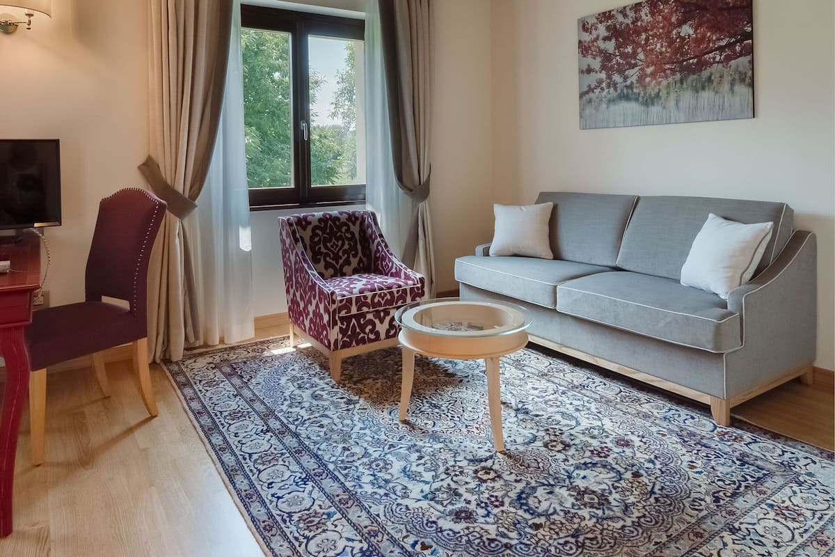 A living room with a wooden ceiling, a grey sofa, patterned armchairs, a red chair, a small round table, and a large window with curtains.