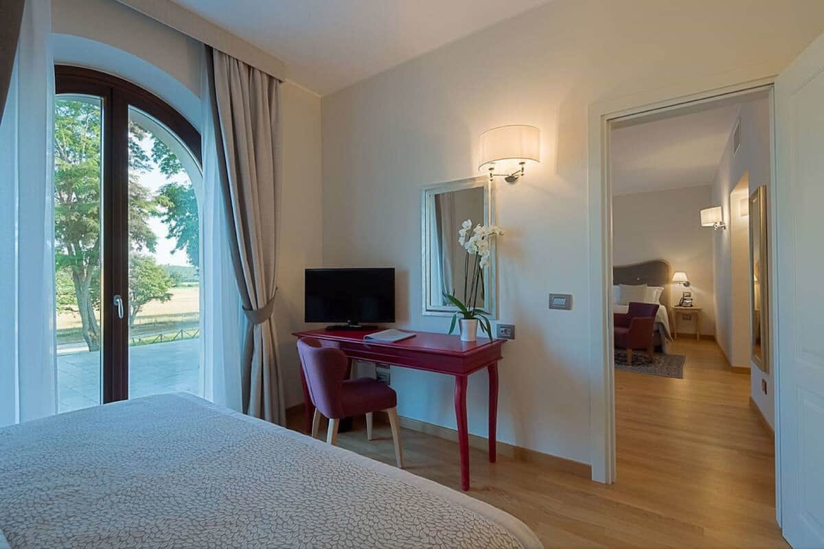 A hotel room with a bed, a desk with a TV, a mirror, and a view of the outdoors.