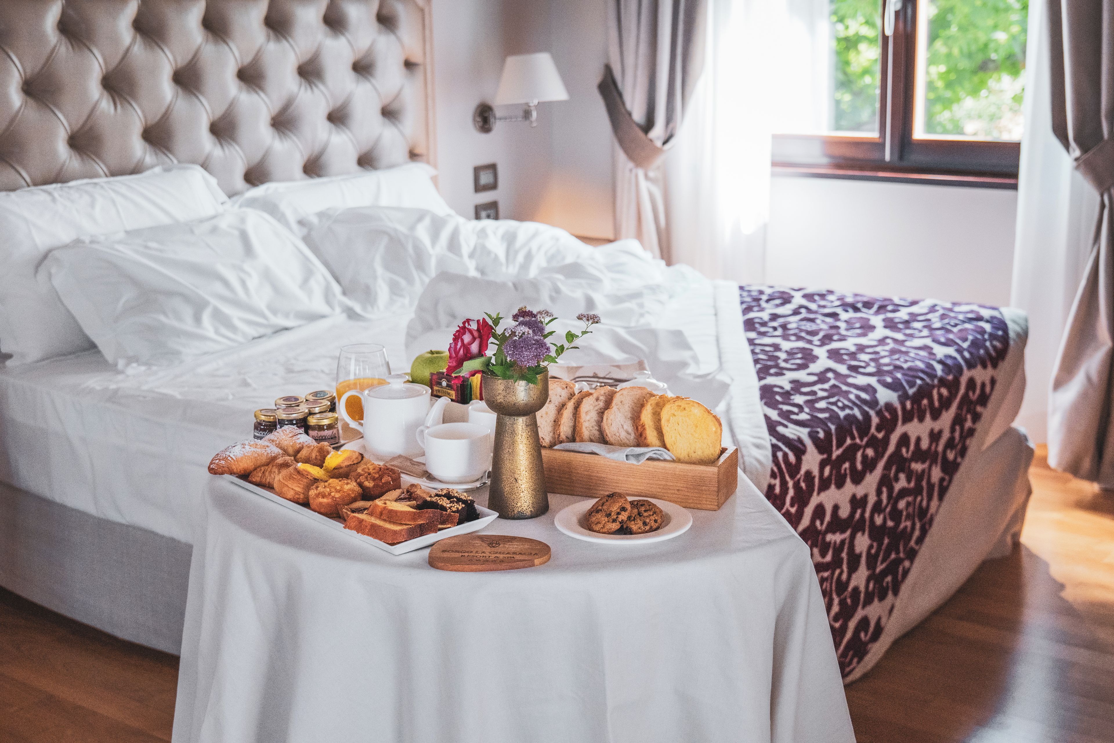 Un lit bien fait, recouvert d’une courtepointe blanche, une assiette de pâtisseries, une cafetière et un vase rempli de fleurs, le tout posé sur une table de chevet.