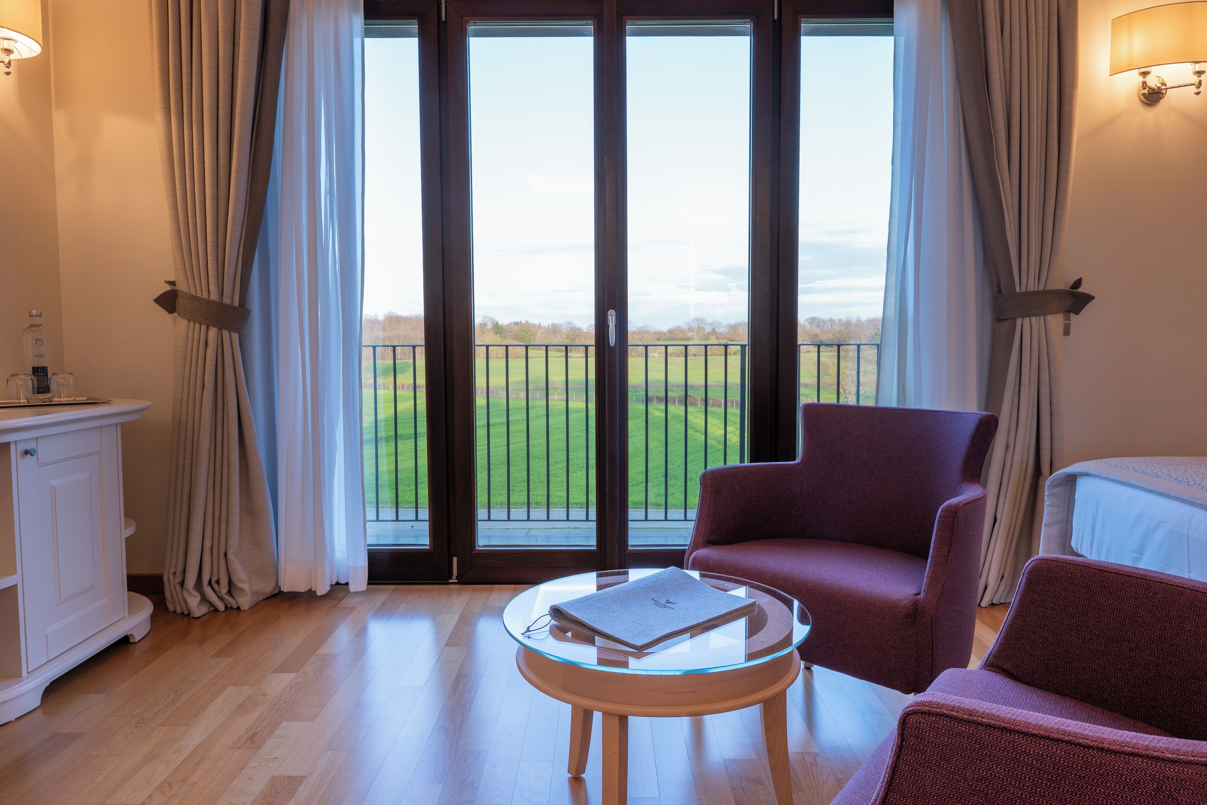 Una habitación con un gran ventanal corredero que da a un balcón, una mesa redonda de vidrio con un libro, un sillón rojo y un armario blanco.