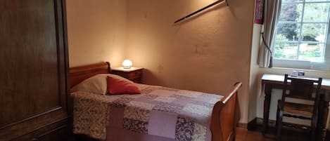 A bedroom with a wooden bed, a nightstand with a lamp, a wooden desk with a chair, and a window with curtains.
