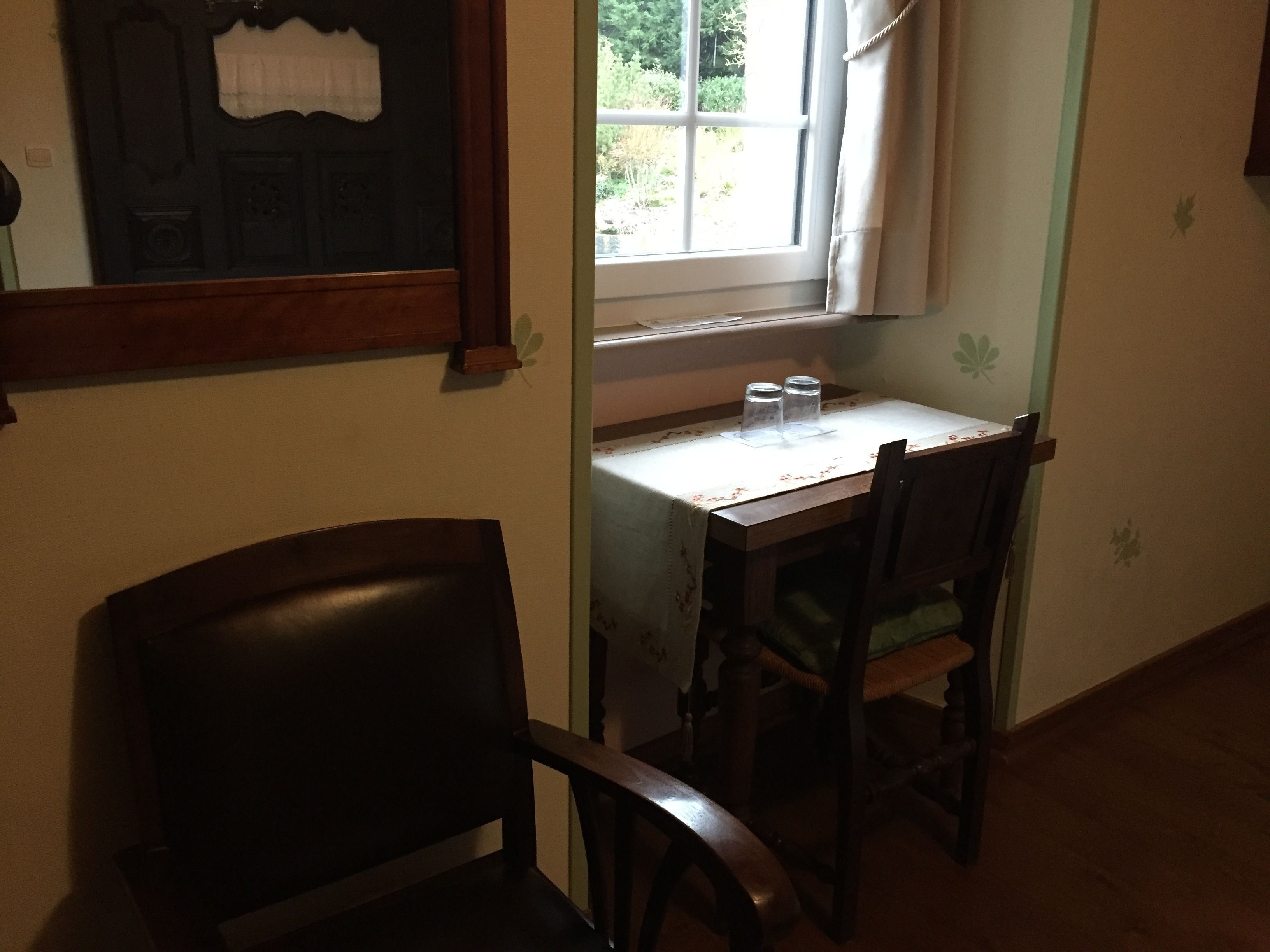 A room with a wooden desk and two chairs, a window with a view of greenery, and a door with a decorative panel.