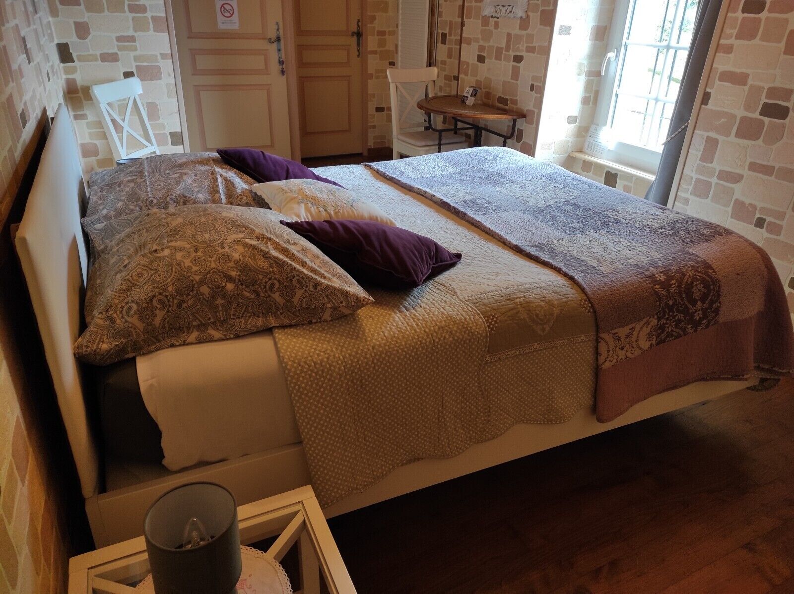 Un llit amb roba de llit estampada, una taula petita amb una espelma i una cadira en una habitació amb parets de maó i una finestra.