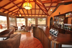A wooden bar area with a curved counter, several chairs, and a view of the outdoors through large windows. - Elephant Rock Luxury Camp Tarangire (Arusha)