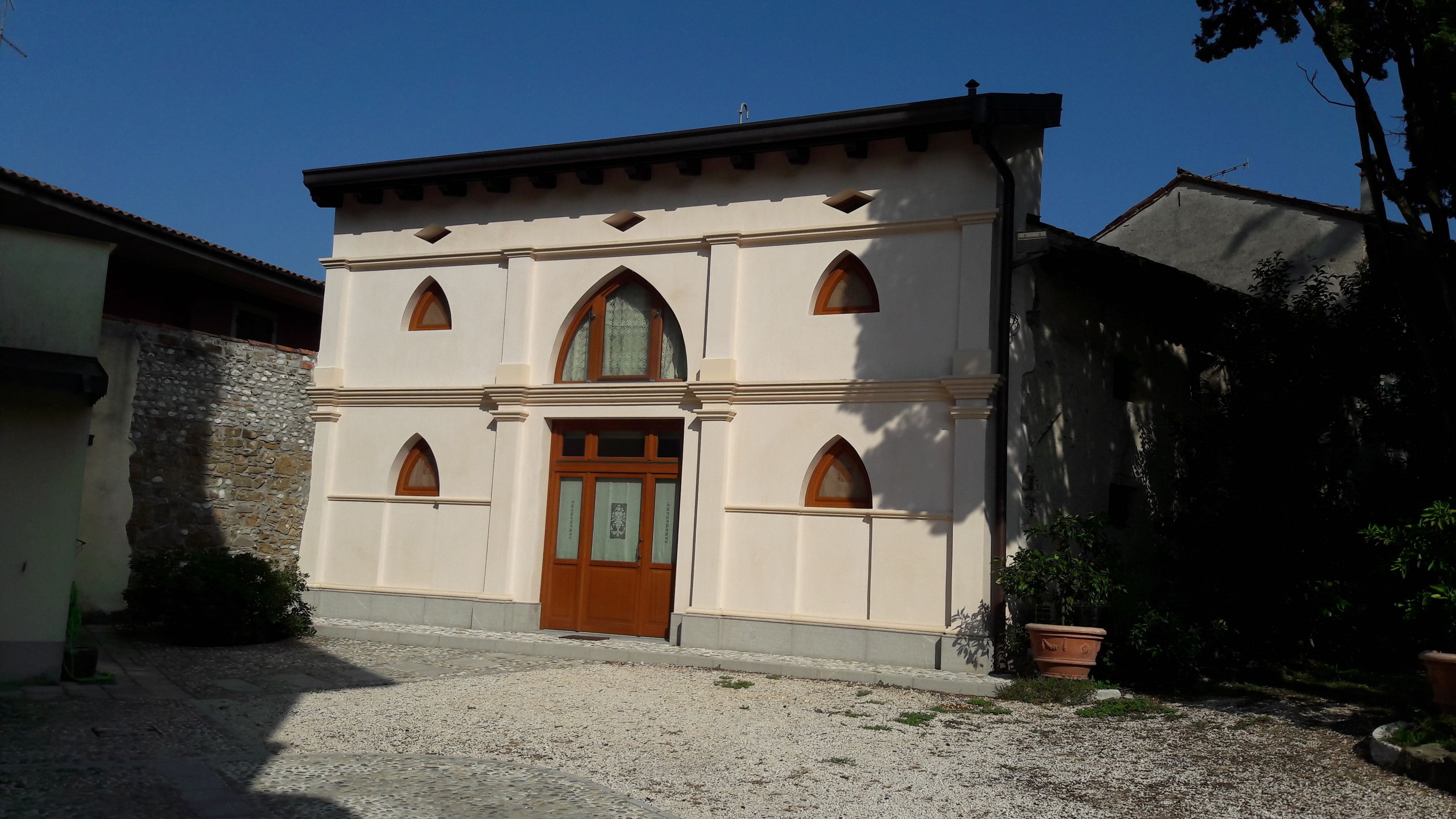 A two-story building with a central entrance, arched windows, and a stone wall to the left.