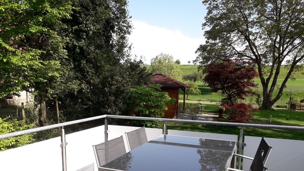 Un balcon doté d’une table en verre et de chaises en métal, donnant sur un jardin agrémenté d’une structure en bois et d’arbres.