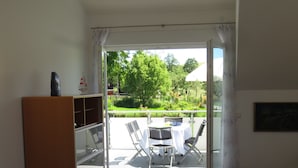A room with a wooden floor, a dining table, a glass-top table, and a view of a garden through a sliding glass door.