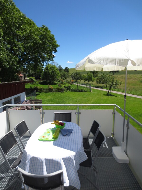 Varanda com mesa posta para refeições, cadeiras e vista para uma paisagem verde com um guarda-sol branco.