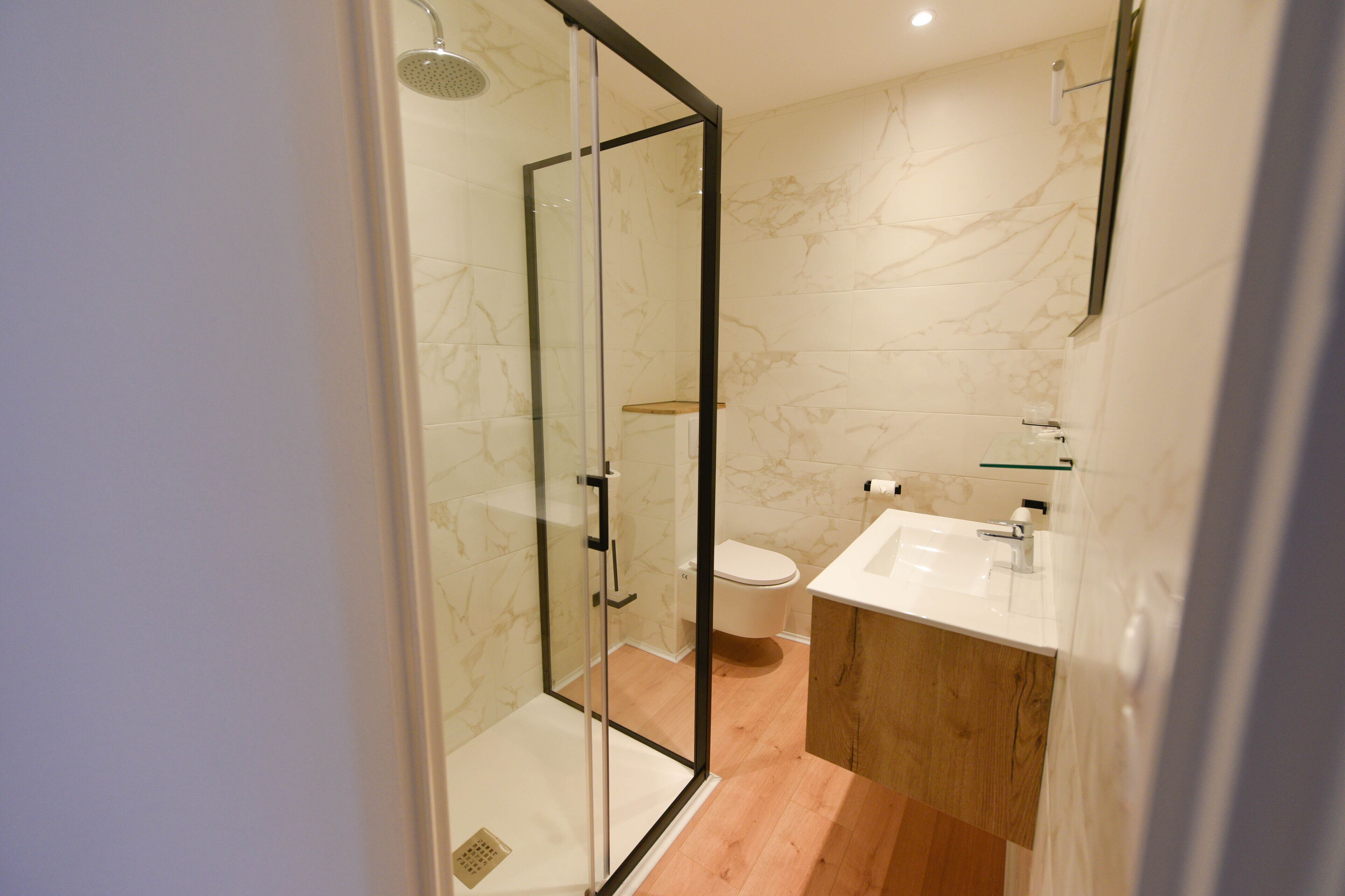 A modern bathroom with a glass-enclosed shower, a wooden vanity, and a marble-tiled wall.