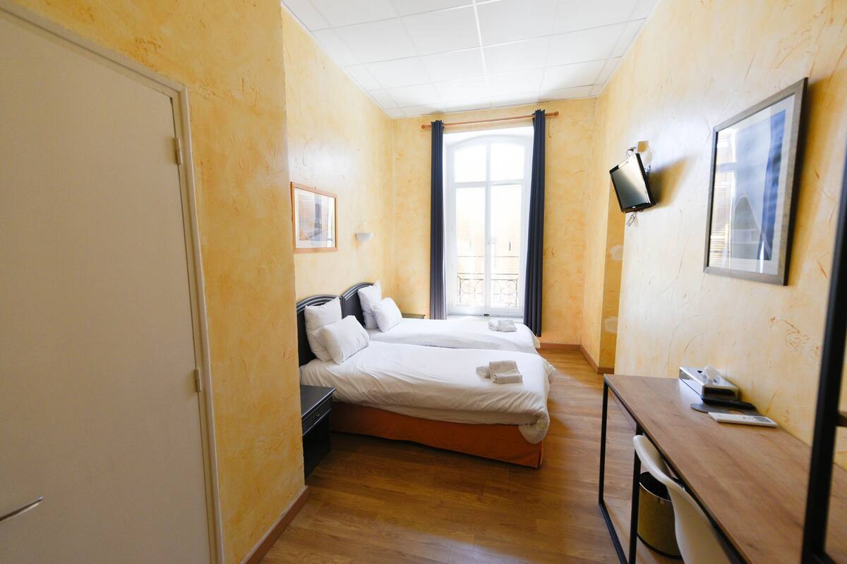 A hotel room with two beds, a desk, a TV, and a window with curtains.