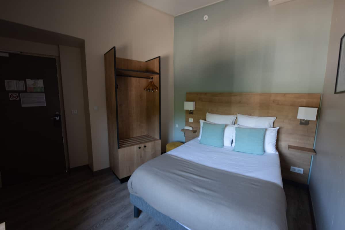 A hotel room with a bed, a wooden wardrobe, a nightstand, and a wall-mounted lamp.