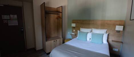 A hotel room with a bed, a wooden wardrobe, a nightstand, and a wall-mounted lamp.