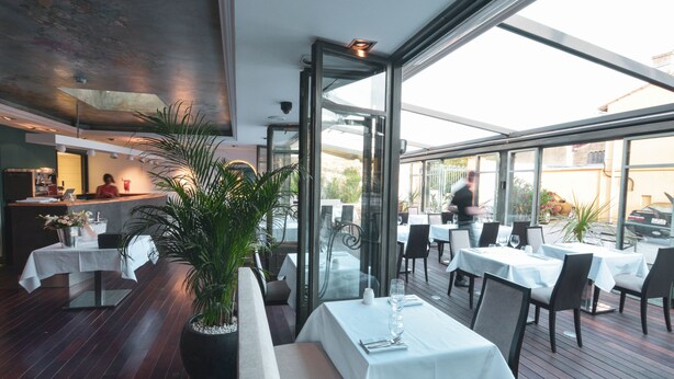 Un espace de restauration en plein air avec des tables pour deux, entouré de plantes en pot et offrant une vue sur la rue.