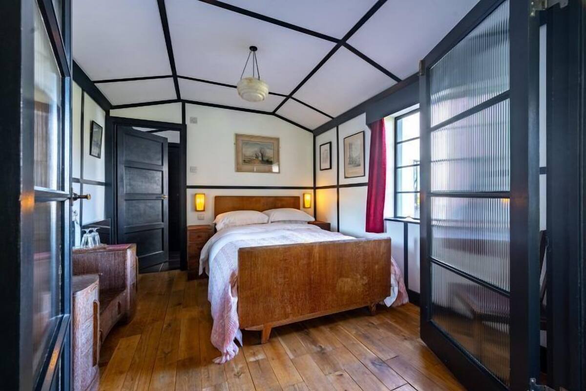 A bedroom with a wooden bed, a nightstand, a dresser, a large window with blinds, and a ceiling with a geometric design.